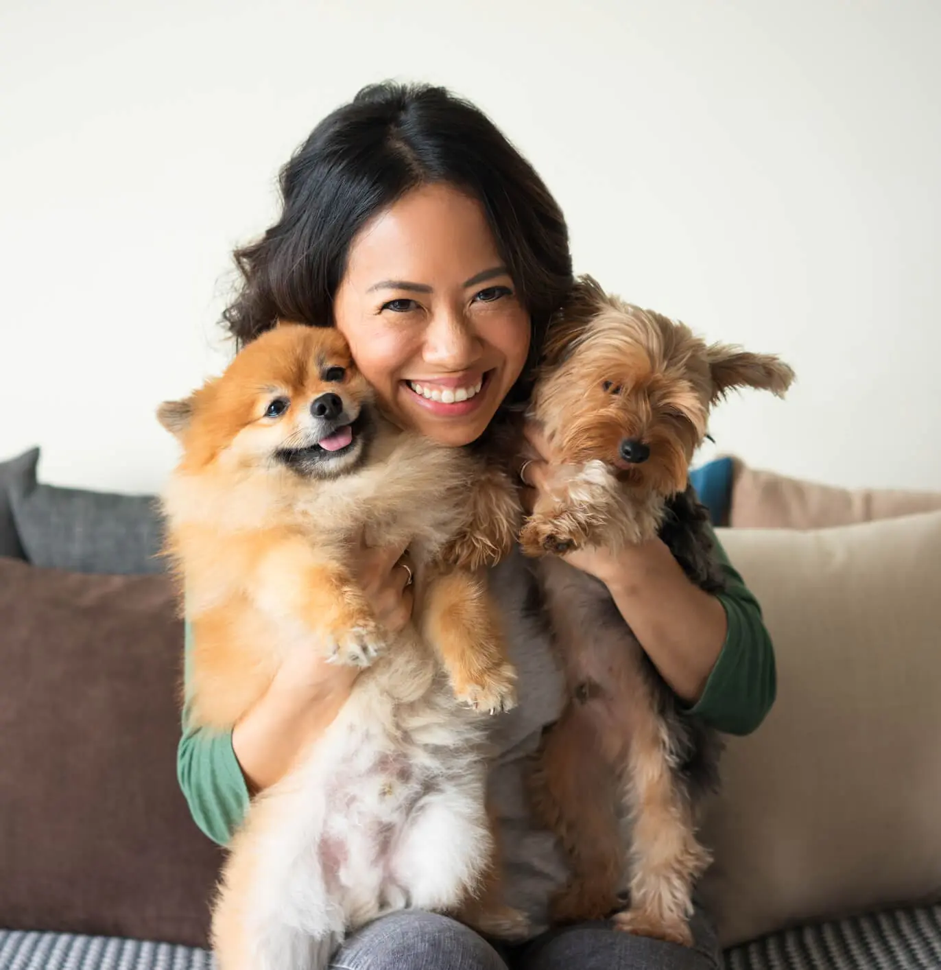 woman smiling while holding two small dogs and sitting on couch indoors